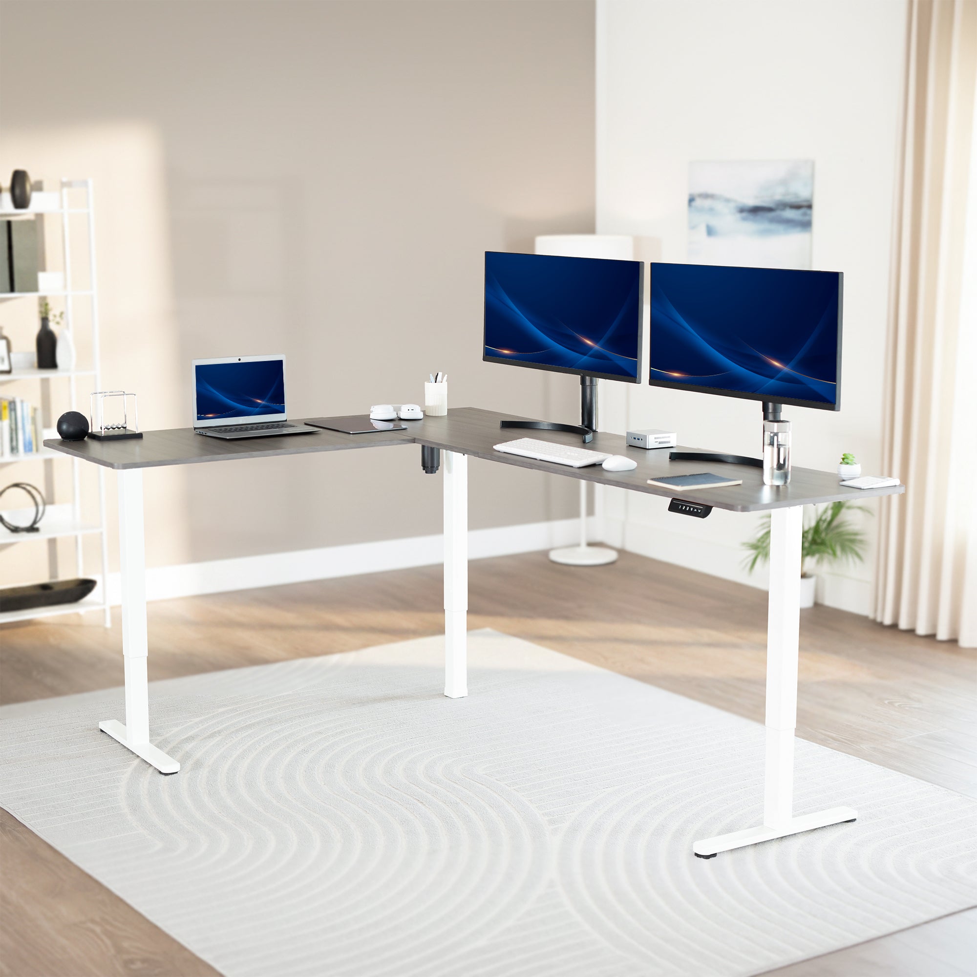 Modern office setup with a standing l-shaped desk, laptop, and two monitors in a bright room.