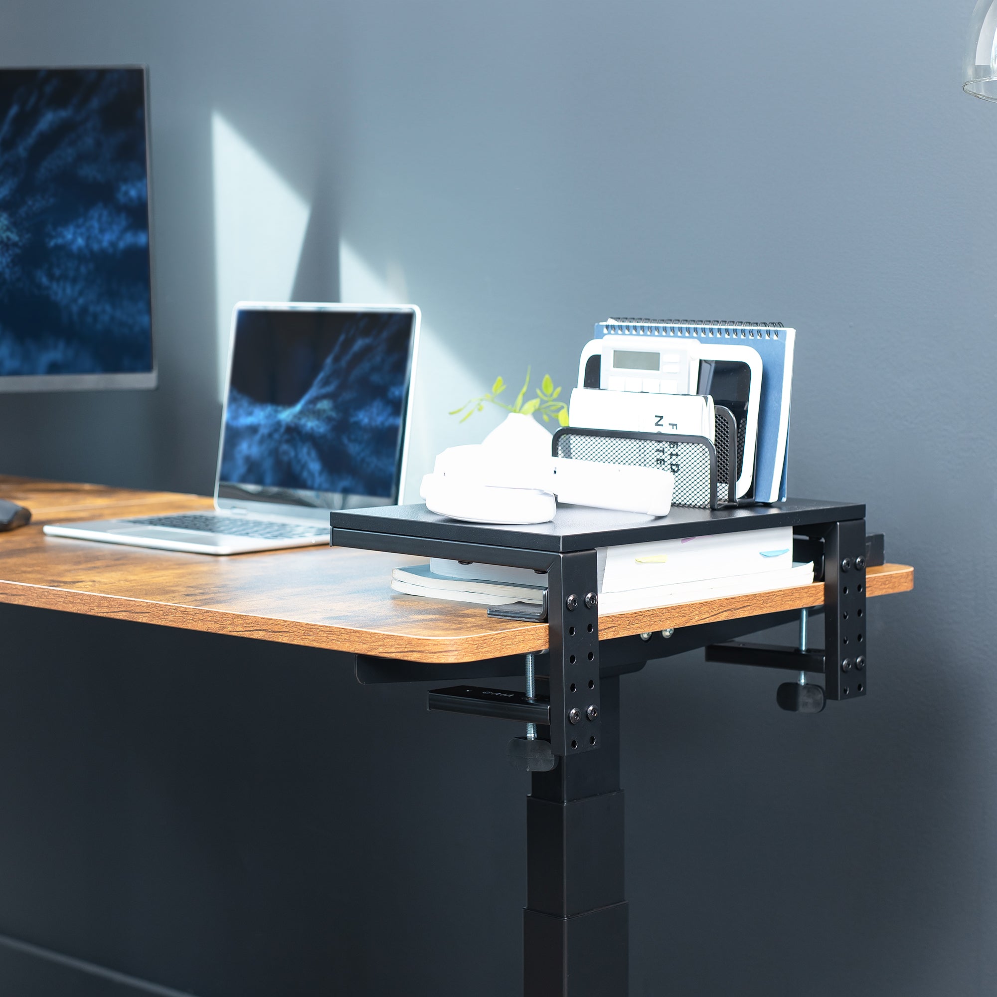 Wooden desk with a black adjustable shelf and office items against a gray wall.