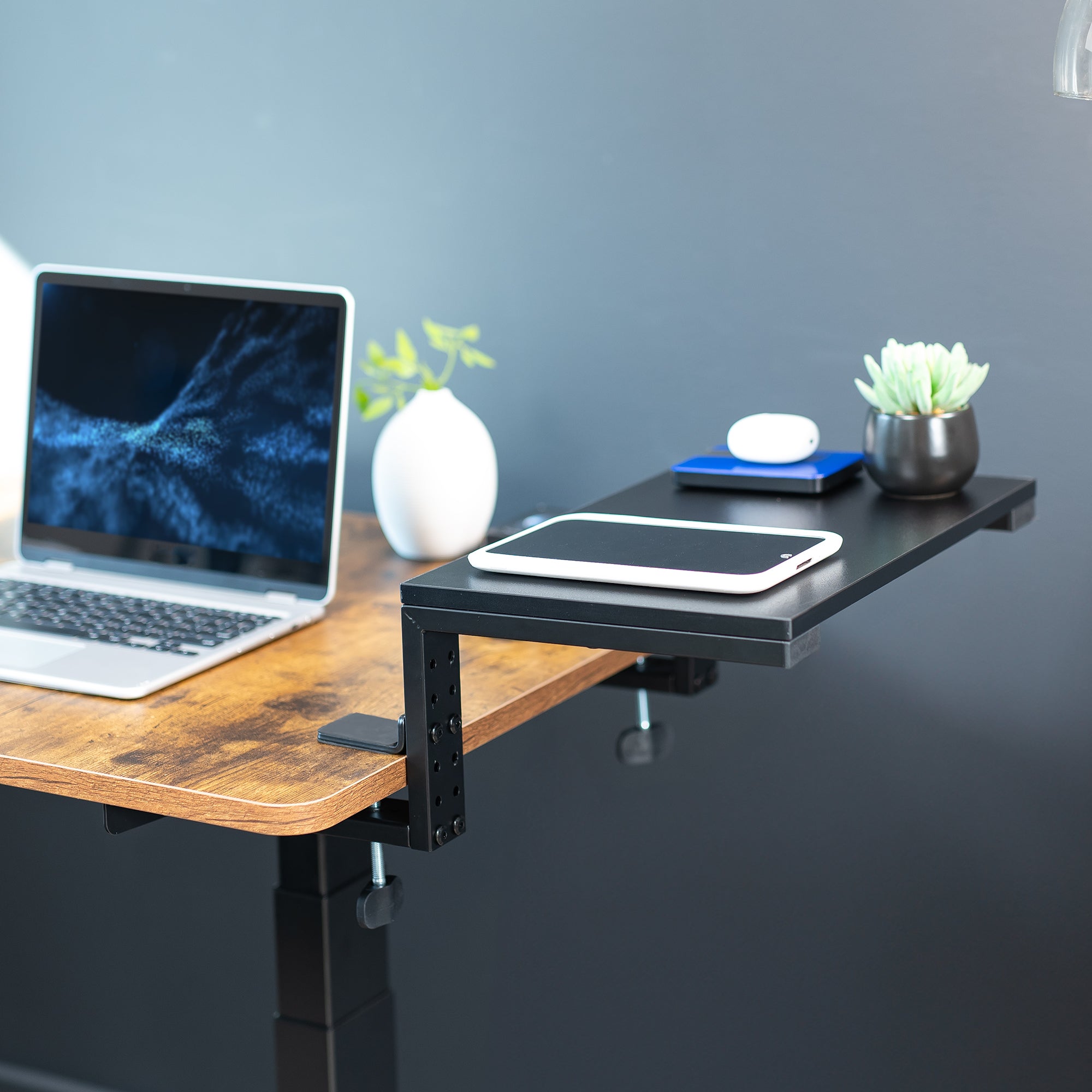 Clamp-on desk extension with laptop and tablet on a wooden desk against a gray wall.