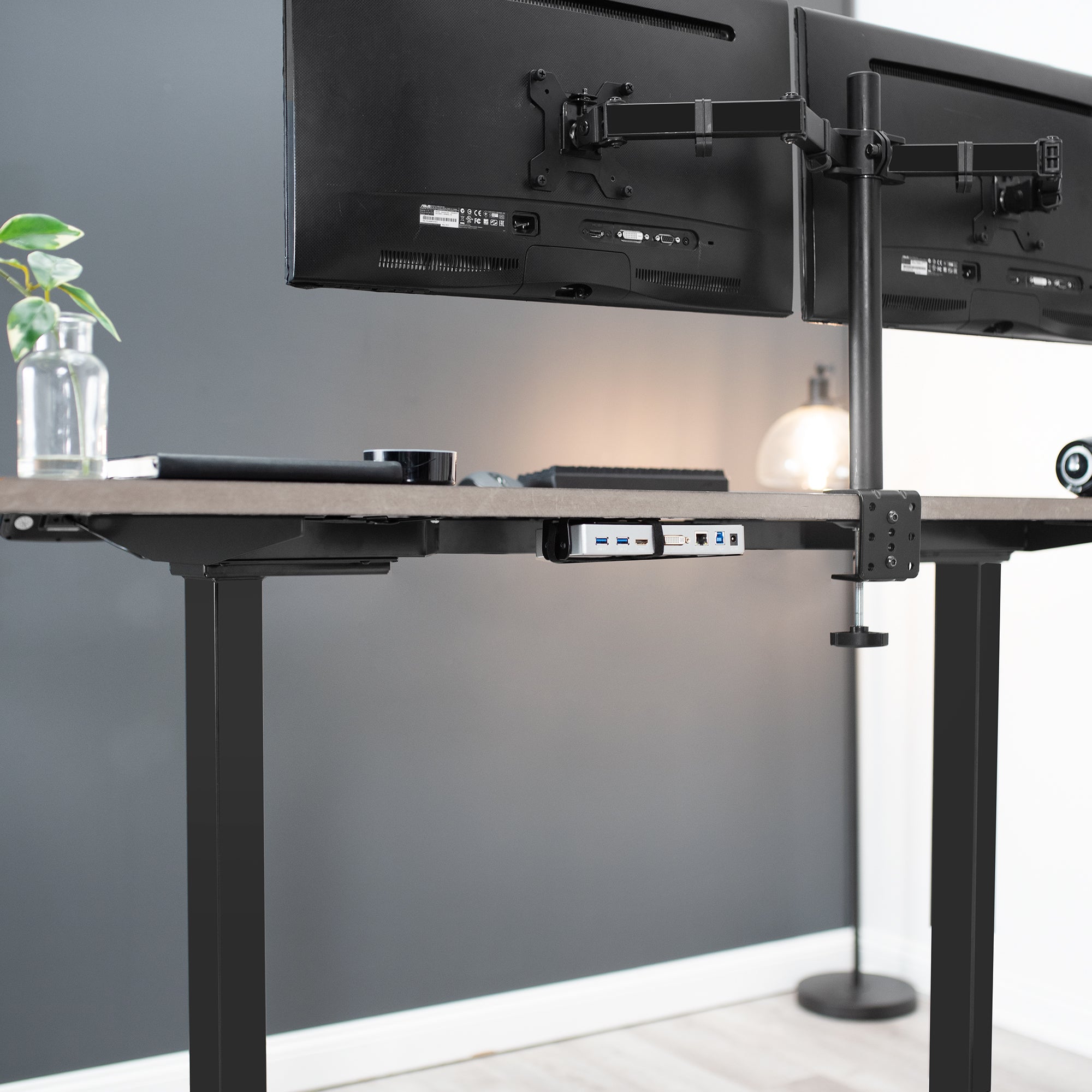 Modern office setup with a adjustable desk, VESA adapter docking station, and dual monitor stand against a gray wall.