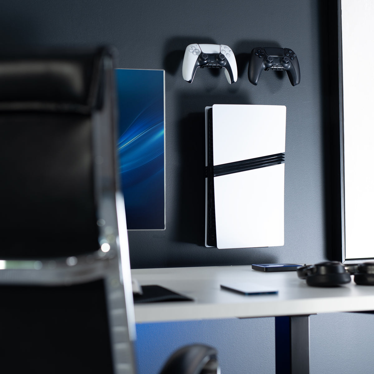A white PlayStation 5 console hangs on a dark gray wall in a modern home office setup, with two wireless controllers mounted above it and a desk visible in the foreground.