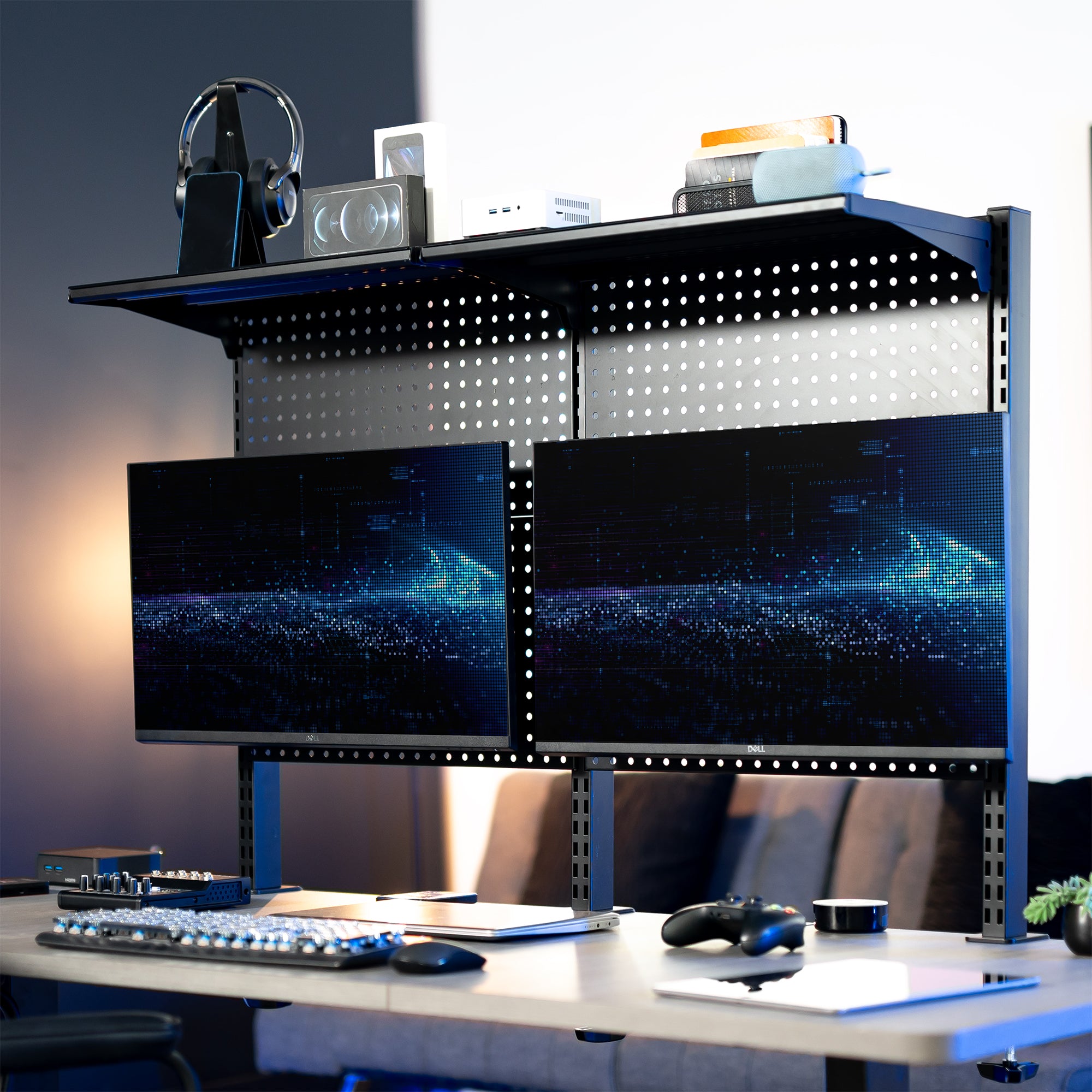 A black clamp-on pegboard monitor mount with a shelf, holding two flat-screen monitors, a speaker, and various desk accessories.