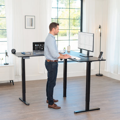 Man using a standing l-shaped desk in a bright room with large windows.