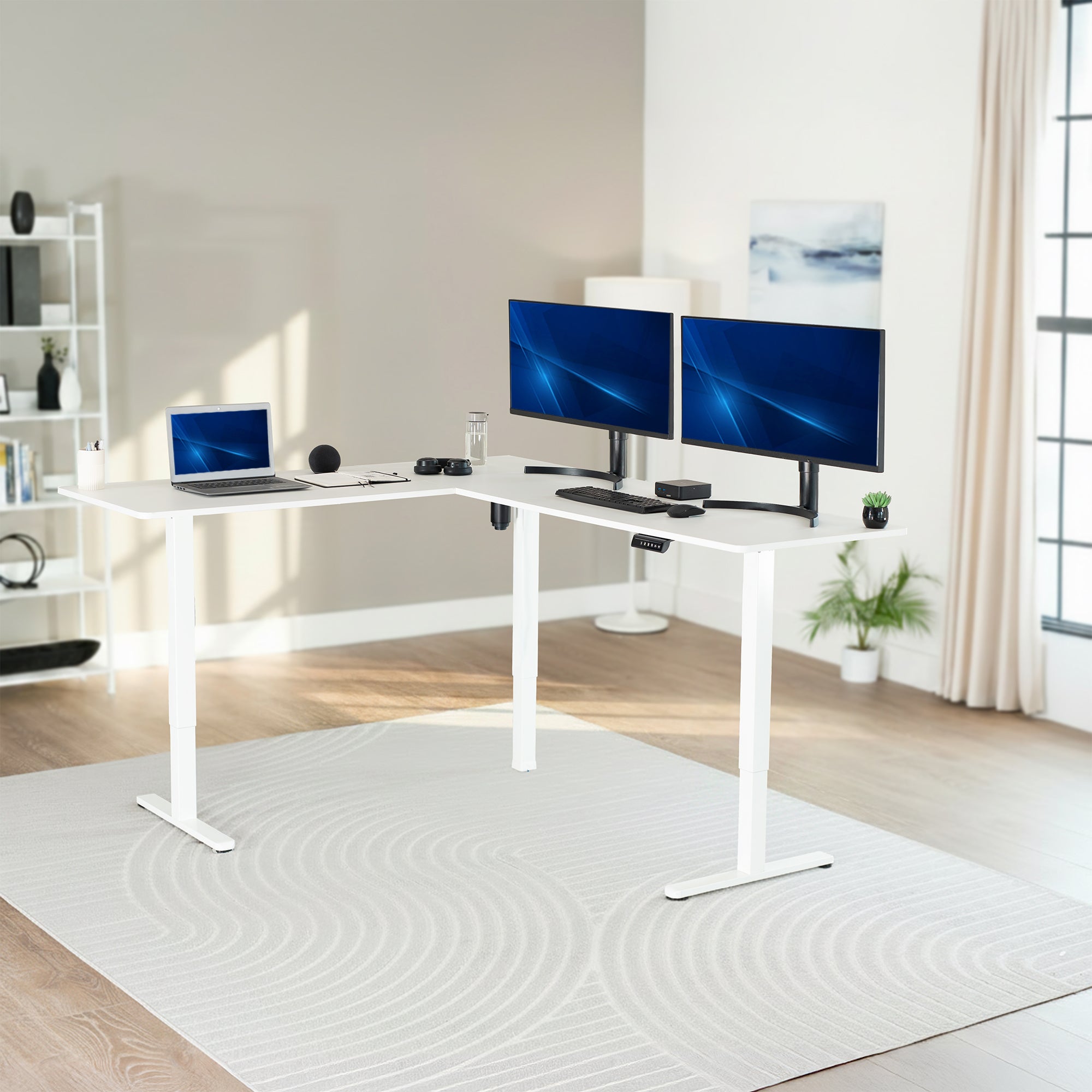 White L-shaped desk with two computer monitors and a laptop in a room with a large window.