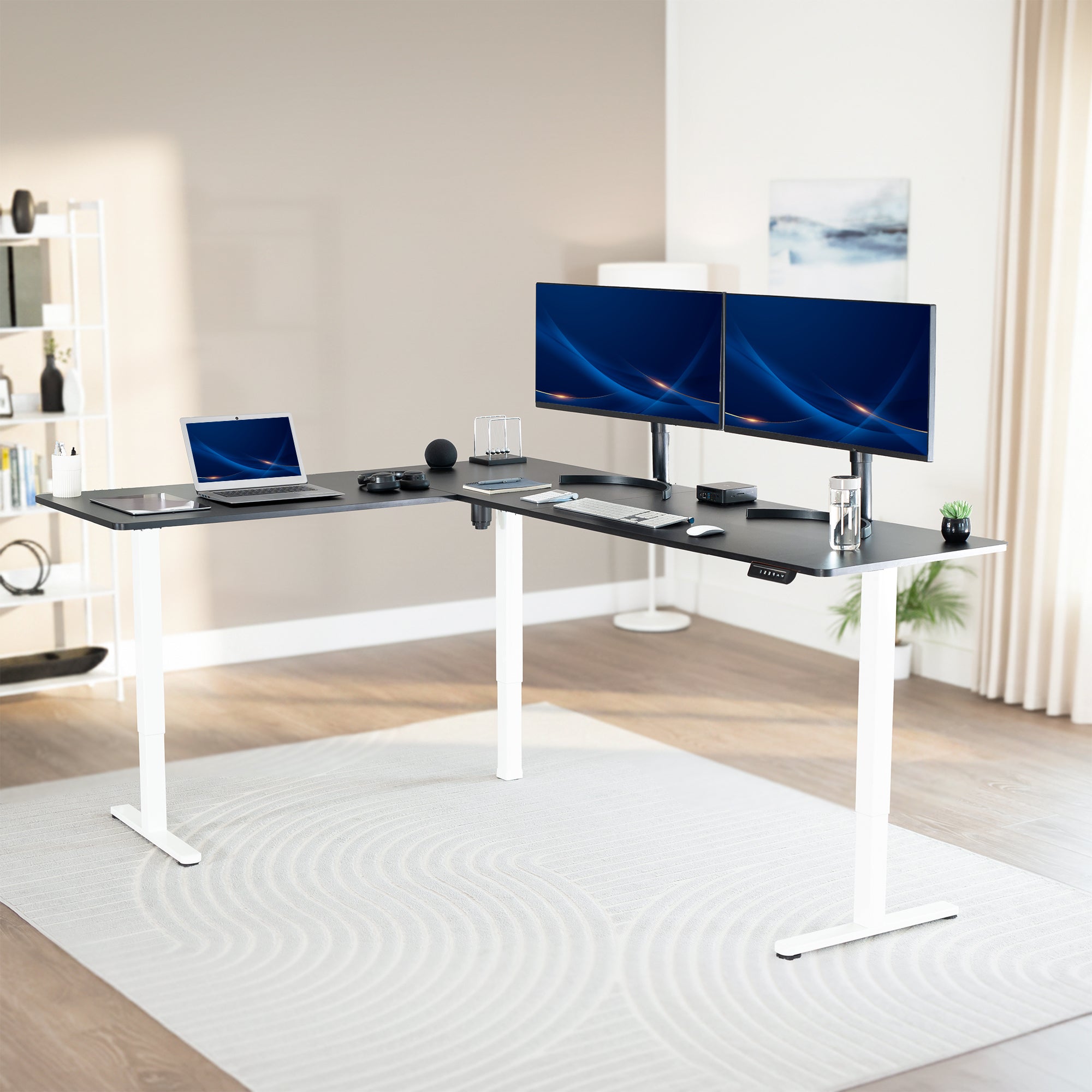 Modern office setup with a standing l-shaped desk, laptop, and two monitors in a bright room.