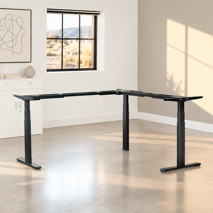 A black adjustable standing desk with a L-shaped frame and T-style legs, shown in a modern home office setting with a window, concrete floor, and beige accent wall.