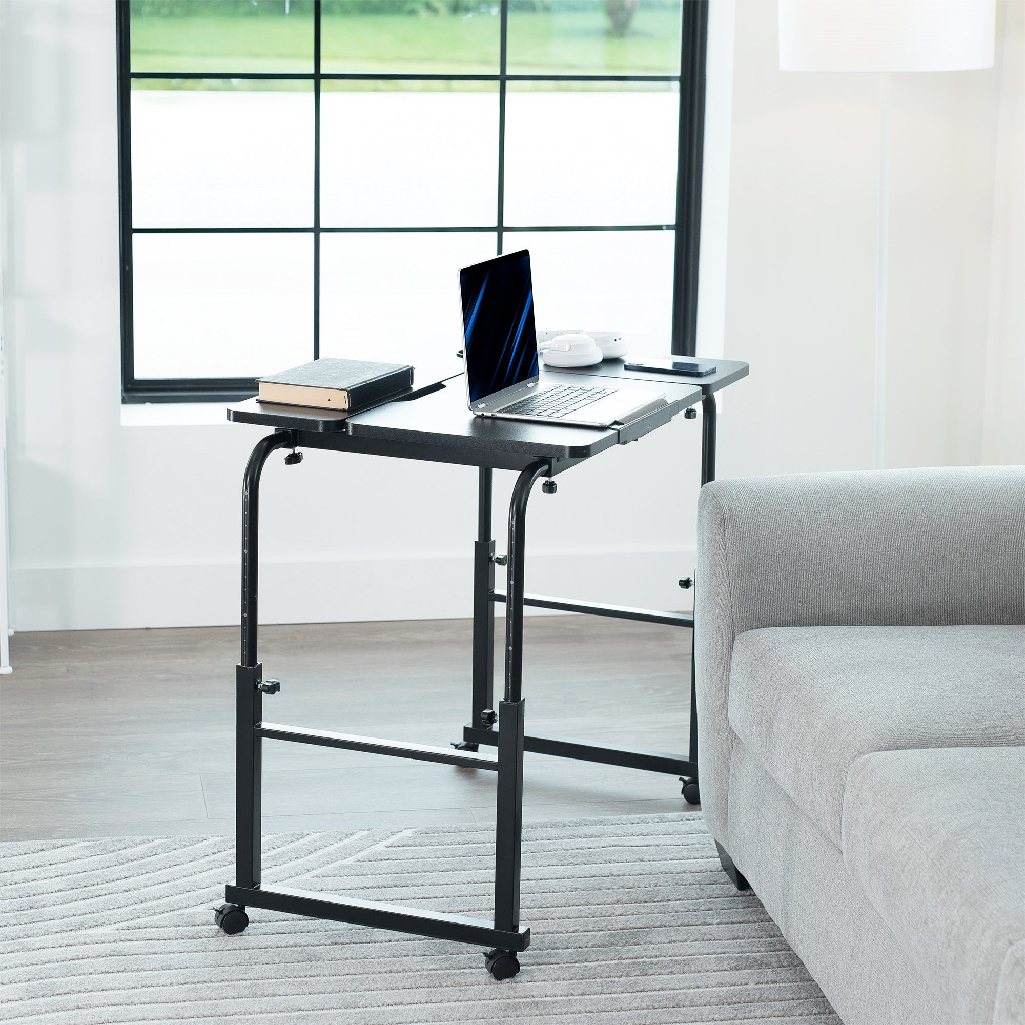 Black adjustable desk in a room with a laptop and books on it, next to a gray sofa.