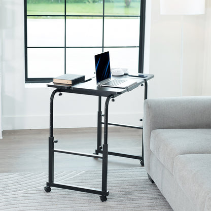 Black adjustable desk in a room with a laptop and books on it, next to a gray sofa.