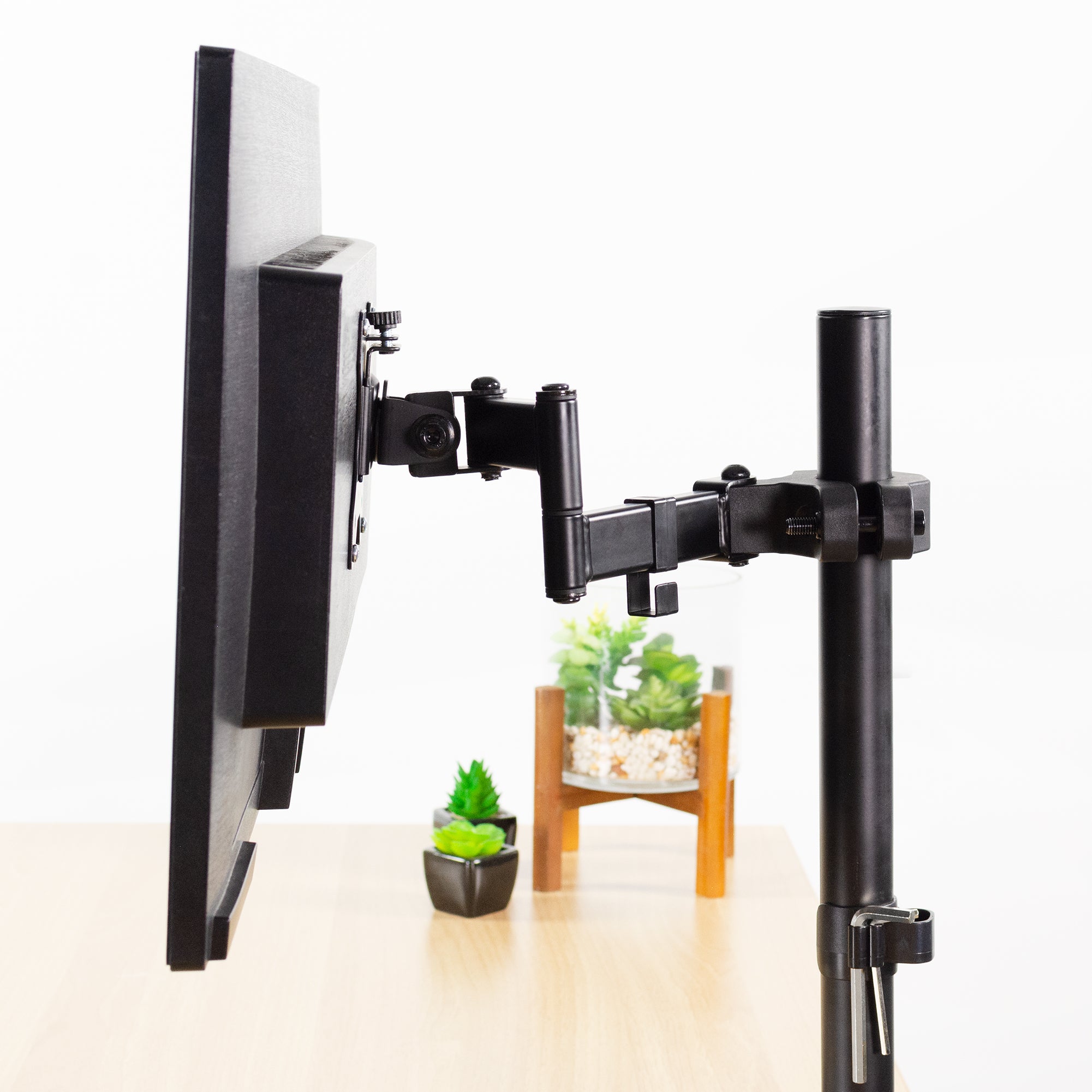 Pole Mount VESA Monitor Arm - 17" to 32" Screens holding a large black screen, mounted on a matching stand over a light wood desk. Small potted plants are visible in the background against a clean white wall.