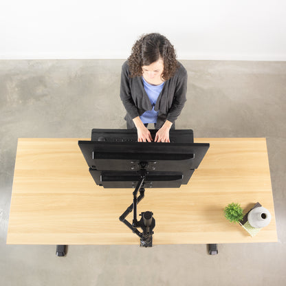 Overhead view of a woman standing at a light wood desk with black computer monitors on an adjustable black stand, in a minimalist room with a concrete floor and white wall.