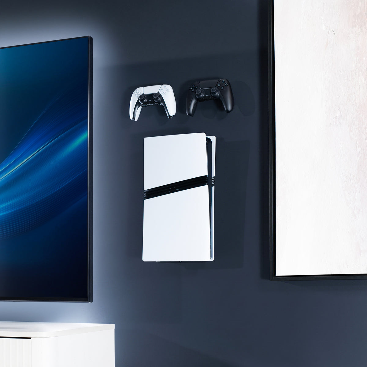 A white PlayStation 5 console with a black accent strip is mounted on a dark wall, flanked by two game controllers, with a TV and framed wall art visible in a modern room setting.
