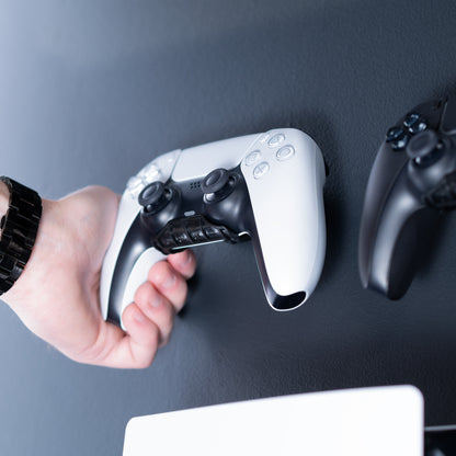 A hand wearing a black watch places a white and black PlayStation 5 DualSense wireless controller on controller hooks against a dark gray wall, with another black controller partially visible on the right.