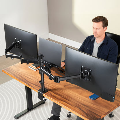 A man sits at a wooden standing desk using dual monitor arm setups with a laptop in between computer monitors and a smartphone in a modern home office setting.